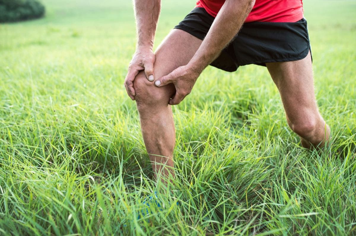 A person in athletic clothing outdoors holding their knee in pain.